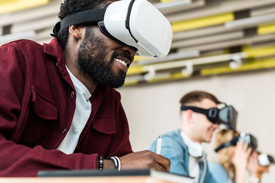 A man sits at a desk with people in the foreground. He is wearing VR goggles and smiling.